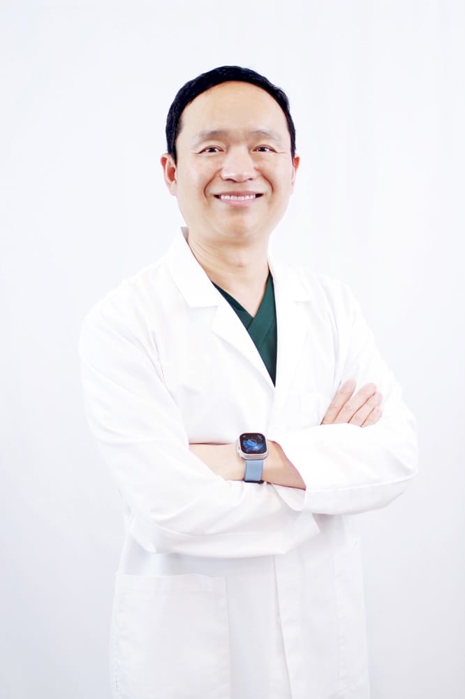 Dr. Hanbit Chi in White Coat – Alki Dental Dr. Hanbit Chi stands confidently with arms crossed, wearing a white lab coat and a smartwatch, smiling against a plain white background – Alki Dental