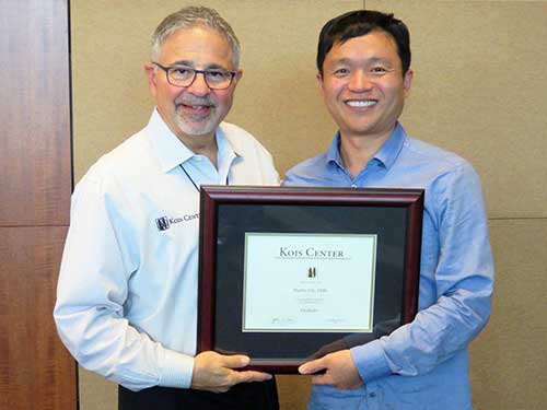 Dr. Hanbit Chi Receiving Recognition – Alki Dental Dr. Hanbit Chi, DDS, wearing a blue long-sleeve shirt, smiles while holding a framed certificate alongside another man in a white shirt with a Kois Center logo – Alki Dental