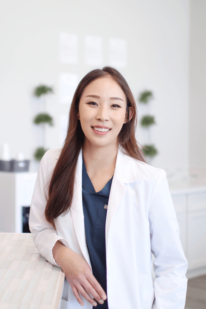 Dr. Sara Park in Clinic – Alki Dental Dr. Sara Park smiles warmly while leaning against a counter, dressed in a white coat over navy scrubs, with a clean and modern dental office background – Alki Dental