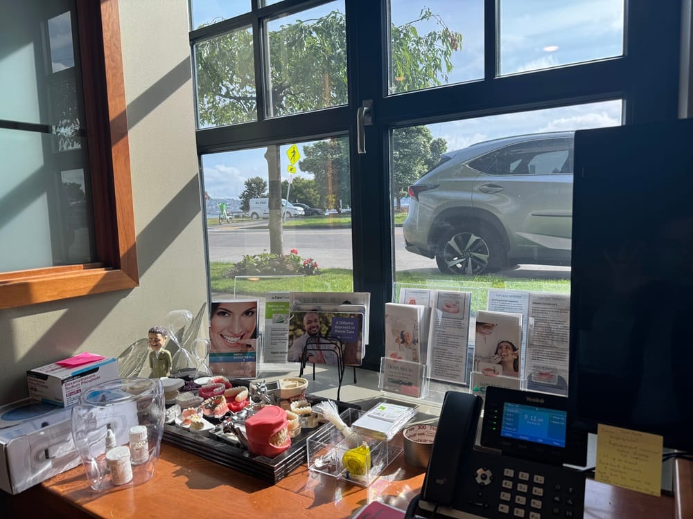 Dental Display by the Window – Alki Dental A sunlit dental office window area displaying dental models, brochures, and informational pamphlets next to a phone and computer monitor – Alki Dental