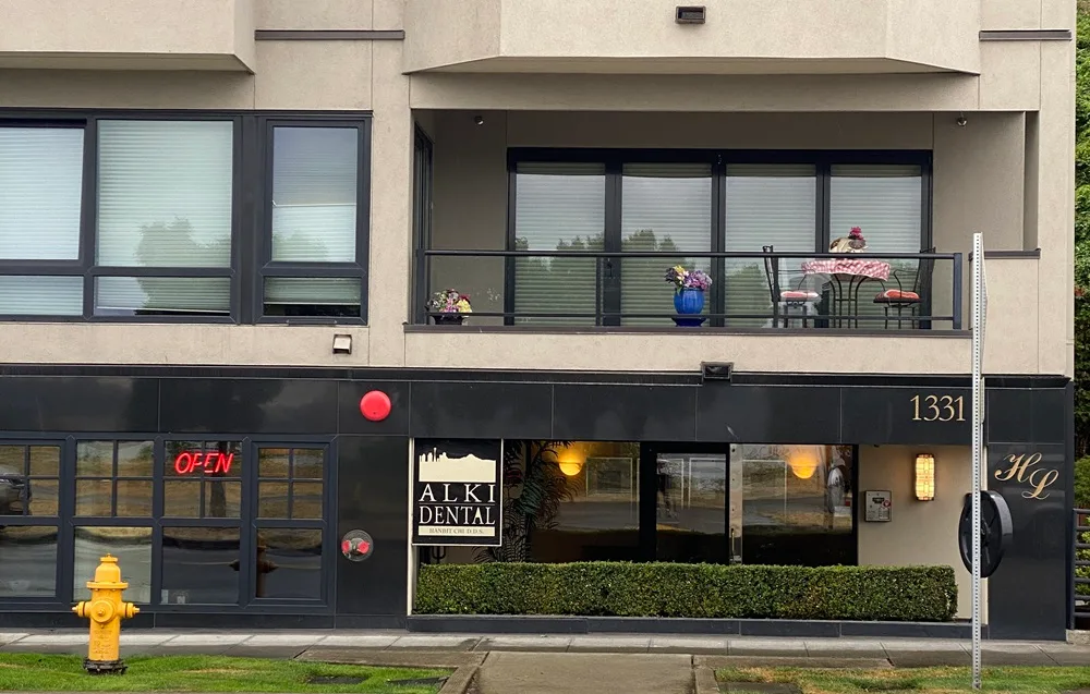The exterior view of Alki Dental with large windows, a neon "OPEN" sign, and a black and beige facade beneath a residential balcony – Alki Dental