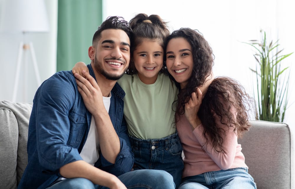 Happy Family on Couch – Alki Dental A smiling family of three sits closely on a couch, with the young daughter between her parents in a bright living room – Alki Dental