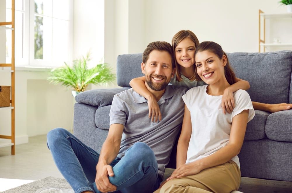 Smiling Family at Home – Alki Dental A happy family of three sits on the floor in front of a gray couch, smiling together in a bright, cozy living room – Alki Dental
