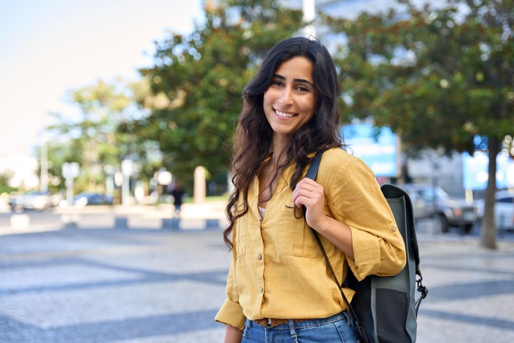Young Woman with Backpack Smiling – Alki Dental A young woman wearing a mustard yellow shirt and jeans smiles while standing outdoors with a backpack – Alki Dental