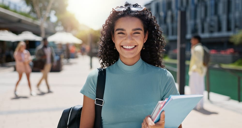 Smiling Student Outdoors – Alki Dental A cheerful young woman with curly hair and a backpack smiles while holding books outside on a sunny day – Alki Dental