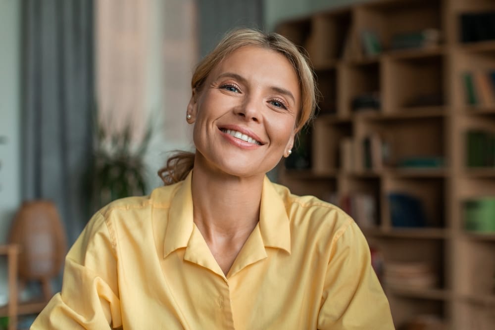 Woman in Yellow Shirt Smiling – Alki Dental A woman in a yellow shirt smiles warmly while sitting in a cozy room with shelves and indoor plants – Alki Dental